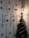 A decorated Christmas tree is situated beside a wall adorned with hanging red ribbons and golden and red baubles. A lit-up pendant lamp casts a warm glow on the decorations, which include small Christmas trees and bells. A Santa Claus ornament is perched on the tree, which is wrapped with colorful tinsel.