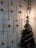 A decorated Christmas tree is situated beside a wall adorned with hanging red ribbons and golden and red baubles. A lit-up pendant lamp casts a warm glow on the decorations, which include small Christmas trees and bells. A Santa Claus ornament is perched on the tree, which is wrapped with colorful tinsel.