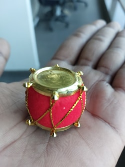 A small, ornate drum with a golden top and bottom, red body, and golden embellishments is being held in the palm of a hand. Background shows a blurry indoor office environment with chairs visible.