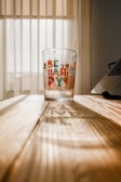 A glass with colorful text and geometric designs sits on a wooden table. Sunlight enters from the window behind, casting shadows on the table’s surface.