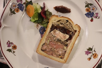 A decorated plate features a slice of pâté en croûte, showcasing a layered interior with various textures and colors, from meats to a smooth filling, encased in a golden crust. It is garnished with a small portion of mixed pickled vegetables and a smear of a dark chutney-like condiment. The plate is adorned with floral designs along the edges.