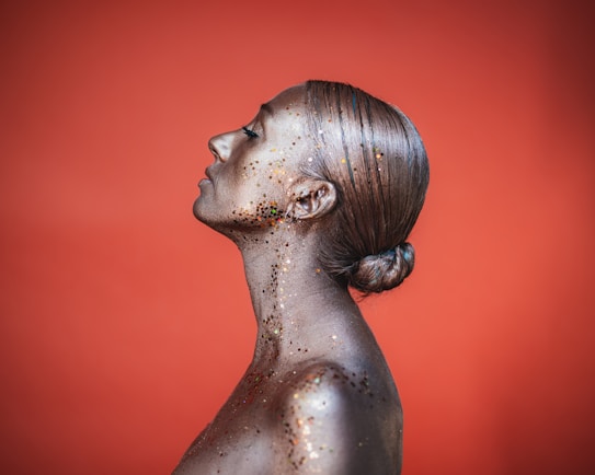 A person with their skin covered in metallic paint and glitter, viewed in profile with a serene expression. The background is a solid, warm orange color, and the hair is styled in a sleek, tight bun.