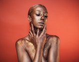 A person is covered in shimmering gold body paint, with a striking pose against a warm, orange background. The individual's eyes are closed, and they gently touch their face and neck with both hands. Sparkling glitter is scattered across the shoulders and upper chest, enhancing the texture and radiance.