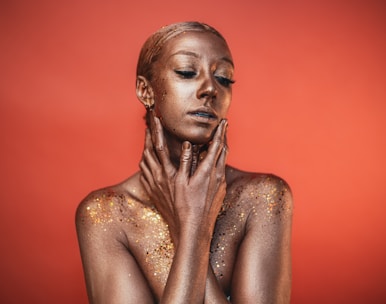 A person is covered in shimmering gold body paint, with a striking pose against a warm, orange background. The individual's eyes are closed, and they gently touch their face and neck with both hands. Sparkling glitter is scattered across the shoulders and upper chest, enhancing the texture and radiance.