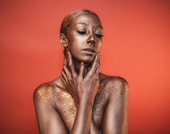 A person is covered in shimmering gold body paint, with a striking pose against a warm, orange background. The individual's eyes are closed, and they gently touch their face and neck with both hands. Sparkling glitter is scattered across the shoulders and upper chest, enhancing the texture and radiance.