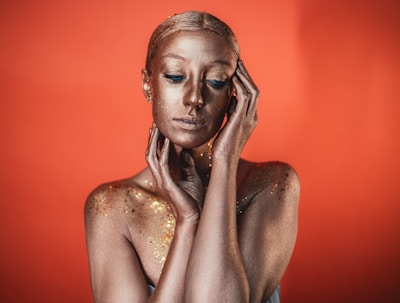 A person is covered in a metallic, gold-like body paint against a vibrant orange background. Their eyes are closed, and their hands are resting gently on their face, creating an artistic and serene composition.