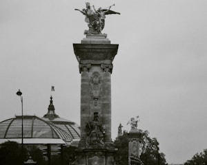 A black and white photograph of a tall stone monument topped with a detailed sculpture of a figure holding a trumpet. Below the sculpture, there is ornate engravings and a seated statue. In the background, a large glass and steel dome can be seen, likely part of a historic building, with trees framing the scene.