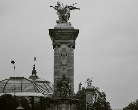 A black and white photograph of a tall stone monument topped with a detailed sculpture of a figure holding a trumpet. Below the sculpture, there is ornate engravings and a seated statue. In the background, a large glass and steel dome can be seen, likely part of a historic building, with trees framing the scene.