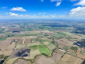 An aerial view of a vast landscape consisting of various agricultural fields in different shades of green and brown. A network of roads and small patches of forests are scattered across the terrain, intersected by winding streams or irrigation channels. The sky is bright blue with scattered clouds casting shadows on the fields.