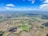 An aerial view of a vast landscape consisting of various agricultural fields in different shades of green and brown. A network of roads and small patches of forests are scattered across the terrain, intersected by winding streams or irrigation channels. The sky is bright blue with scattered clouds casting shadows on the fields.