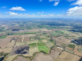 An aerial view of a vast landscape consisting of various agricultural fields in different shades of green and brown. A network of roads and small patches of forests are scattered across the terrain, intersected by winding streams or irrigation channels. The sky is bright blue with scattered clouds casting shadows on the fields.