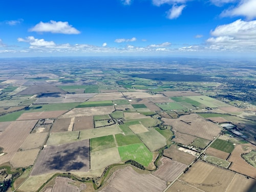 An aerial view of a vast landscape consisting of various agricultural fields in different shades of green and brown. A network of roads and small patches of forests are scattered across the terrain, intersected by winding streams or irrigation channels. The sky is bright blue with scattered clouds casting shadows on the fields.