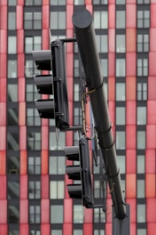 A close-up view of a traffic light system mounted on a pole set against a high-rise building exterior with alternating red and black panels. The traffic lights are black and appear to be in an off state, with visible wires and metal attachments.