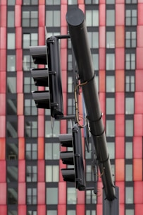 A close-up view of a traffic light system mounted on a pole set against a high-rise building exterior with alternating red and black panels. The traffic lights are black and appear to be in an off state, with visible wires and metal attachments.