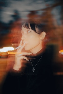 A person with a contemplative expression holds a cigarette, surrounded by a blurred, warm-toned background that creates an ethereal effect. The soft light highlights the person's facial features, jewelry, and the glow from the cigarette.