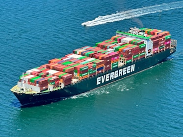 A large cargo ship with containers stacked high on its deck cruises through the ocean. The ship is labeled 'Evergreen' in bold white letters. A smaller boat is visible in the distance creating a trail in the water.