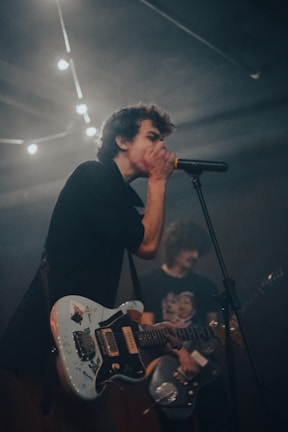 A musician passionately sings into a microphone while playing an electric guitar, with another person in the background also holding a guitar. The lighting is dim, and there are string lights visible above, creating a moody atmosphere.