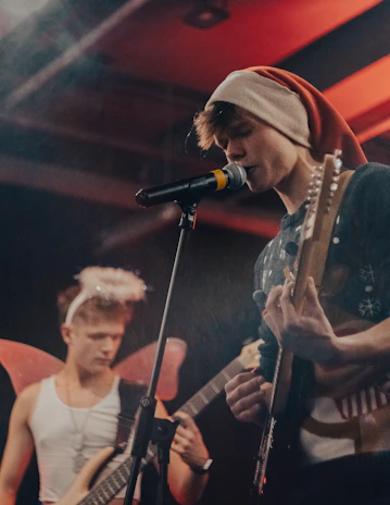 A young musician with a Santa hat is singing into a microphone while playing an electric guitar. Another person in the background is playing a bass guitar and wearing angel wings and a halo headband. The setting is dimly lit with a warm color tone.