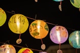 A series of colorful paper lanterns are hanging in the night sky, each decorated with unique designs and handwritten characters. The lanterns are glowing warmly, illuminated from within, creating a vibrant contrast against the dark background.