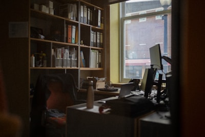 A cozy office space featuring a cluttered desk with multiple monitors, a chair, and various office supplies. A window provides a view of a brick building outside. Shelves filled with binders and books line the wall, adding to the organized yet busy atmosphere.