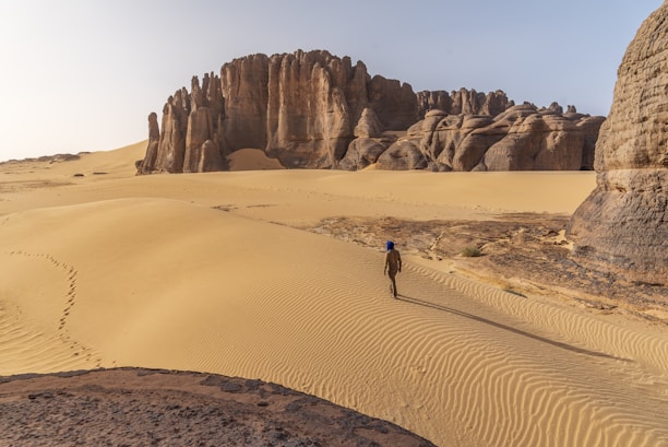 A vast desert landscape featuring towering sandstone formations and expansive sand dunes. A solitary person wearing a blue headscarf walks across the undulating sand, leaving footprints behind.