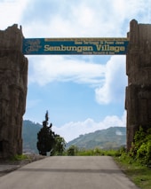 An entrance gate with the words 'Sembungan Village' written on it, flanked by two large stone pillars. The background includes a clear blue sky with some clouds and terraced green hills. A paved path leads through the gate.