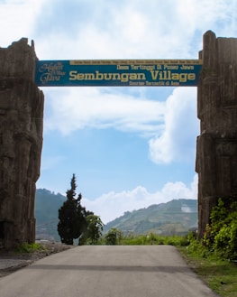 An entrance gate with the words 'Sembungan Village' written on it, flanked by two large stone pillars. The background includes a clear blue sky with some clouds and terraced green hills. A paved path leads through the gate.