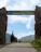 An entrance gate with the words 'Sembungan Village' written on it, flanked by two large stone pillars. The background includes a clear blue sky with some clouds and terraced green hills. A paved path leads through the gate.