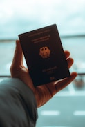 A hand is holding a German passport, which displays the emblem of the country. The background is blurred, possibly an airport or travel location, with a soft focus on the passport itself.