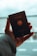A hand is holding a German passport, which displays the emblem of the country. The background is blurred, possibly an airport or travel location, with a soft focus on the passport itself.