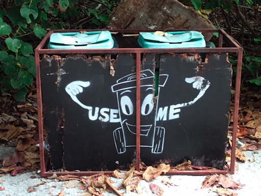 A weathered garbage bin with a cartoon-like illustration on the front, featuring a smiling trash can character pointing to itself with the words 'USE ME' written beside it. The bin is positioned outdoors among fallen leaves and surrounded by green foliage.