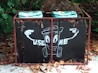 A weathered garbage bin with a cartoon-like illustration on the front, featuring a smiling trash can character pointing to itself with the words 'USE ME' written beside it. The bin is positioned outdoors among fallen leaves and surrounded by green foliage.