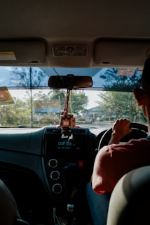 A person is driving a car, viewed from the backseat, with a focus on the interior dashboard. The dashboard features a display showing the time and date. Several small objects, including a USB drive, are hanging below the rearview mirror. Sunlight is streaming through the front windshield, casting shadows and creating reflections.