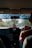 A person is driving a car, viewed from the backseat, with a focus on the interior dashboard. The dashboard features a display showing the time and date. Several small objects, including a USB drive, are hanging below the rearview mirror. Sunlight is streaming through the front windshield, casting shadows and creating reflections.