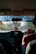 A person is driving a car, viewed from the backseat, with a focus on the interior dashboard. The dashboard features a display showing the time and date. Several small objects, including a USB drive, are hanging below the rearview mirror. Sunlight is streaming through the front windshield, casting shadows and creating reflections.