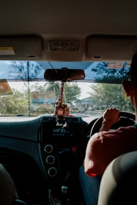 A person is driving a car, viewed from the backseat, with a focus on the interior dashboard. The dashboard features a display showing the time and date. Several small objects, including a USB drive, are hanging below the rearview mirror. Sunlight is streaming through the front windshield, casting shadows and creating reflections.