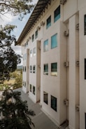 A modern multi-story building with a geometric, minimalist design. The exterior wall is painted in a light beige color with numerous rectangular windows. Air conditioning units are mounted beneath each window. In the foreground, lush green trees add a natural element to the scene and partially obscure the view. The sky is partially visible, showcasing a clear, sunny day.