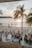 A bustling beachside scene at sunset with people dining at tables on a wooden deck under palm trees. Several groups are sitting and conversing, while others stroll along the sandy shore. Decorative banners hang above the crowd, and the sun sets over the ocean, creating a warm glow on the water.