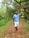 A person stands on a wooded path surrounded by lush greenery, wearing a blue t-shirt, white shorts, and red shoes. They are looking up at a sign hanging from a tree branch. The sign reads, 'The clean air here is a therapy to your lungs. Take a deep breath.'
