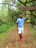 A person stands on a wooded path surrounded by lush greenery, wearing a blue t-shirt, white shorts, and red shoes. They are looking up at a sign hanging from a tree branch. The sign reads, 'The clean air here is a therapy to your lungs. Take a deep breath.'