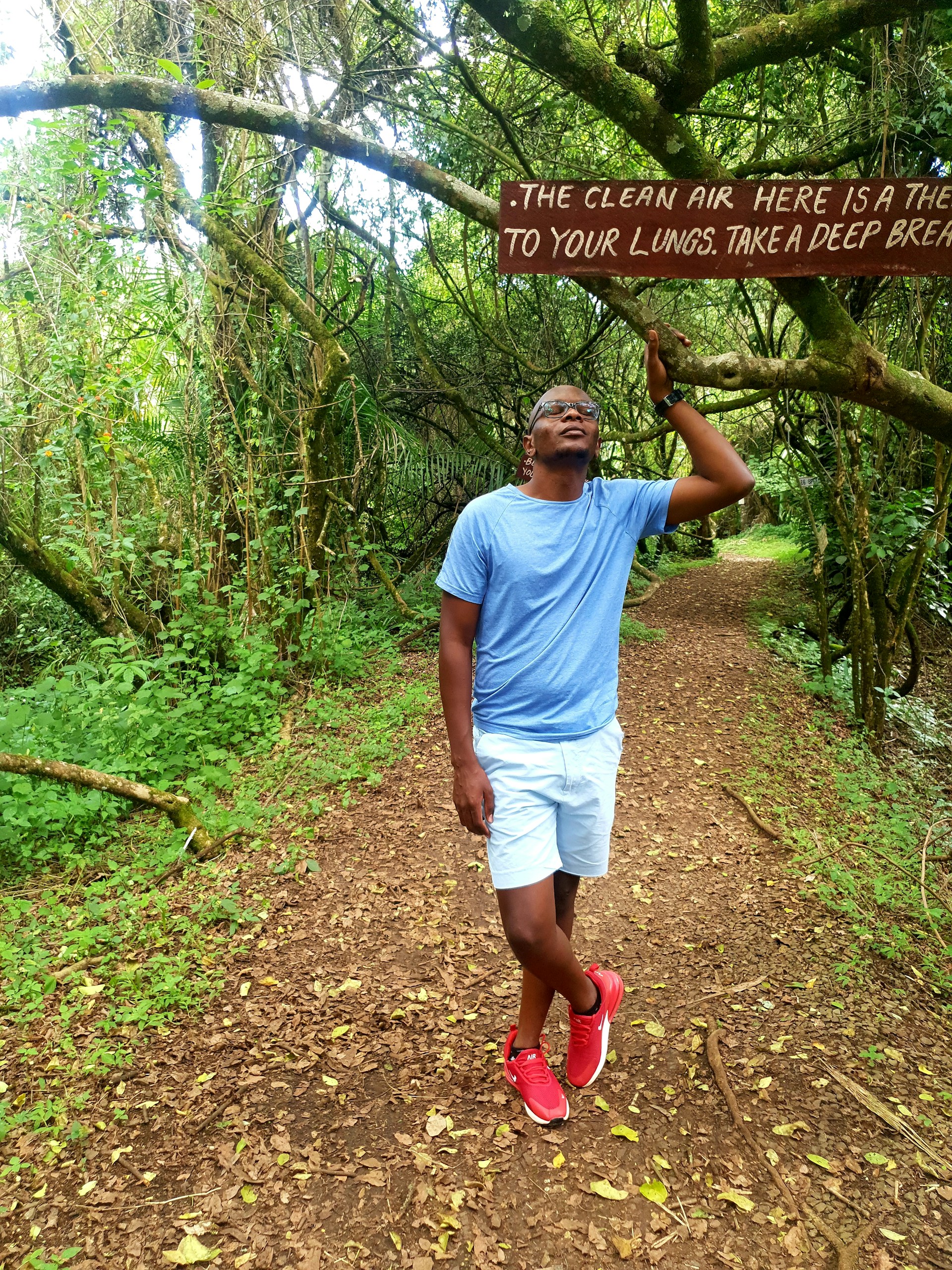 A person stands on a wooded path surrounded by lush greenery, wearing a blue t-shirt, white shorts, and red shoes. They are looking up at a sign hanging from a tree branch. The sign reads, 'The clean air here is a therapy to your lungs. Take a deep breath.'