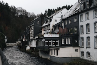 A picturesque row of traditional German-style houses lines a small river, with steep, forested hills in the background. The buildings feature dark timber framing, slate roofs, and large windows. One building displays a sign indicating it is a restaurant. The overcast sky and bare trees suggest a chilly, early winter atmosphere.