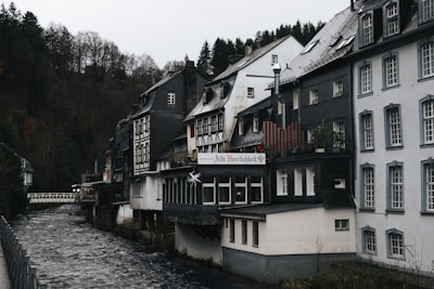 A picturesque row of traditional German-style houses lines a small river, with steep, forested hills in the background. The buildings feature dark timber framing, slate roofs, and large windows. One building displays a sign indicating it is a restaurant. The overcast sky and bare trees suggest a chilly, early winter atmosphere.