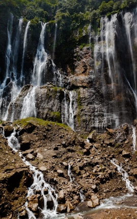 A majestic waterfall cascades down a rugged cliff face covered in lush greenery. The water flows over numerous rock ledges, creating multiple streams and a misty atmosphere. The area is surrounded by dense forest vegetation.
