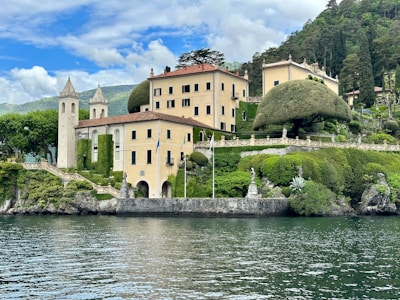 A large, picturesque villa situated on the shore of a lake, surrounded by lush greenery and manicured gardens. The buildings have a warm, light yellow color with dark green shutters. There is a stone tower structure with pointed roofs. Sculptures, flags, and ornate fencing decorate the landscape. The sky is partly cloudy, and the lake water is calm and reflective.