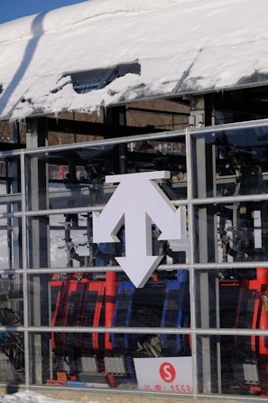 A snow-covered structure with a prominent white multi-directional arrow sign on its glass surface stands in front of a ski lift. Various mechanical components and colorful chairlift seats in red and blue are visible inside the glass enclosure. A small red 'S' symbol is printed on a sign with a few numbers below.