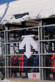 A snow-covered structure with a prominent white multi-directional arrow sign on its glass surface stands in front of a ski lift. Various mechanical components and colorful chairlift seats in red and blue are visible inside the glass enclosure. A small red 'S' symbol is printed on a sign with a few numbers below.