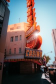 A vibrant city scene featuring a tall, illuminated casino sign with retro-style neon lights. The sign dominates the building facade with the words 'CASINO' and 'HOTEL'. Sunlight peeks through, creating a dramatic lens flare effect. The street is lively, with pedestrians casually walking and a hint of urban hustle.