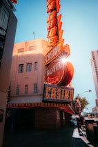 A vibrant city scene featuring a tall, illuminated casino sign with retro-style neon lights. The sign dominates the building facade with the words 'CASINO' and 'HOTEL'. Sunlight peeks through, creating a dramatic lens flare effect. The street is lively, with pedestrians casually walking and a hint of urban hustle.