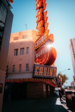 A vibrant city scene featuring a tall, illuminated casino sign with retro-style neon lights. The sign dominates the building facade with the words 'CASINO' and 'HOTEL'. Sunlight peeks through, creating a dramatic lens flare effect. The street is lively, with pedestrians casually walking and a hint of urban hustle.